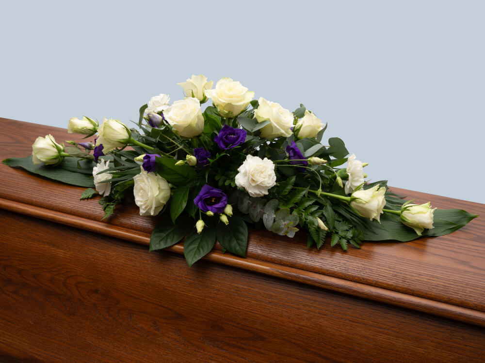 Arkkulaite, jossa valkoisia ruusuja ja violettia eustomaa, vihreät suuren lehdet alla, tammiarkun päällä. Hautauspalvelu Kaiho Oy - Hautajaiskukat kuva