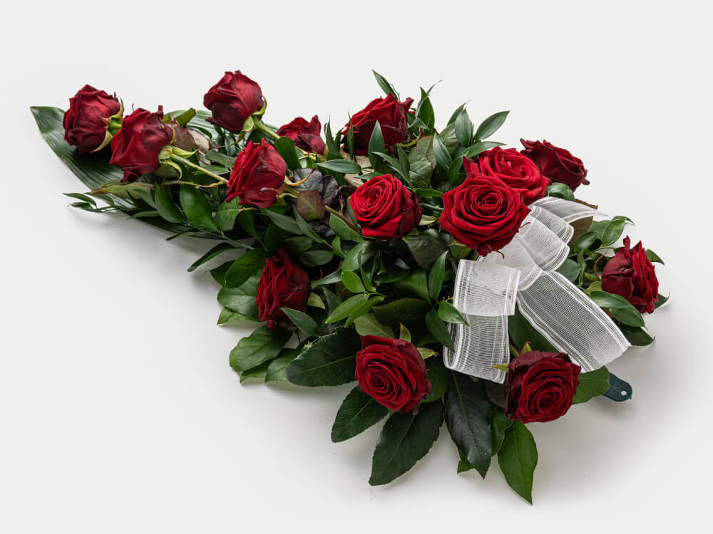 Surulaite, jossa on pisaran muotoon laitettu punaisia ruusuja, vihreitä lehtiä välissä, valkoinen läpinäkyvä nauha ja vihreä kahva pilkistää vähän alta. Hautauspalvelu Kaiho Oy - Hautajaiskukat kuva