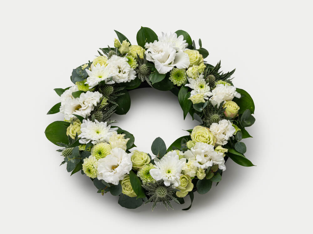 Kukiltaan valkoinen ja vaaleanvihreä pienehkö seppele, alla tummanvihreitä lehtiä. Hautauspalvelu Kaiho Oy - Hautajaiskukat kuva