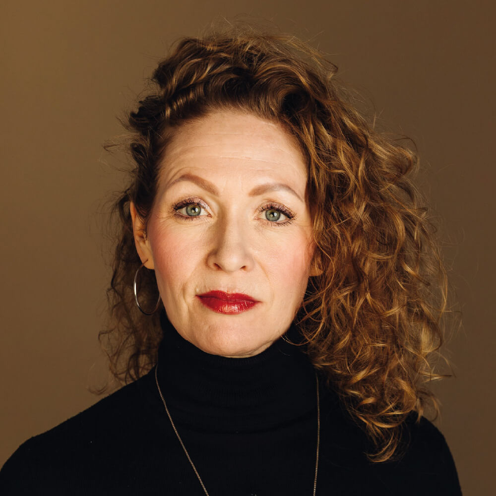 Kiharatukkainen nainen, Sanna Salovuori, katsoo suoraan kameraan. Hautauspalvelu Kaiho Oy - Musiikki ja Puhe kuva